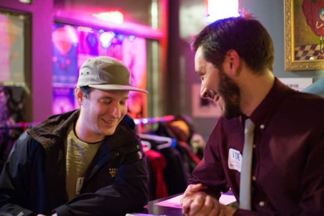 Two men engaged in conversation at a table during the Love Bomb 2017 event, with colorful lighting and a festive atmosphere in the background.